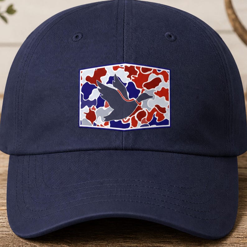 K&ouml;nnte beinhalten: Marineblaue Baseballkappe mit einem rechteckigen Aufn&auml;her. Der Aufn&auml;her zeigt eine Entensilhouette vor einem Tarnmuster in Rot, Wei&szlig; und Blau. Die Kappe ist aus weichem Material und hat einen gebogenen Schirm.