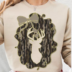 Puede incluir: Sudadera beige con un dise&ntilde;o de silueta de cabeza de ciervo dentro de una corona de camuflaje. La corona tiene un contorno de purpurina dorada y un lazo en la parte superior. El dise&ntilde;o est&aacute; centrado en la parte delantera.