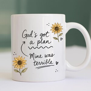 Puede incluir: Una taza de cer&aacute;mica blanca con el texto impreso en negro God's got a plan, Mine was terrible, acompa&ntilde;ada de dos ilustraciones de girasoles amarillos con tallos y hojas verdes, junto con peque&ntilde;os corazones y puntos negros decorativos.