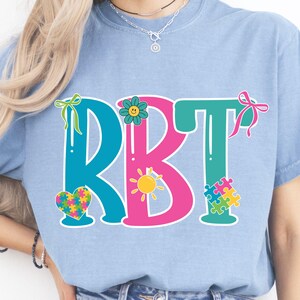 May include: Light blue t-shirt with the letters RBT in teal and pink, decorated with a flower, a sun, a heart, and puzzle pieces. The shirt has a crew neck and short sleeves.