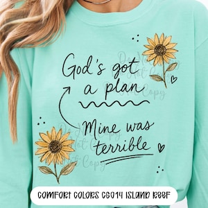 Puede incluir: Una sudadera de manga larga color verde menta con el texto negro God's got a plan, y una flecha que apunta al texto Mine was terrible. El dise&ntilde;o incluye dos girasoles amarillos con hojas verdes, peque&ntilde;os puntos negros y dos corazones.