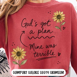 Puede incluir: Una sudadera de cuello redondo color carmes&iacute; con el texto impreso en negro God's got a plan, Mine was terrible, acompa&ntilde;ada de dos girasoles amarillos con tallos y hojas verdes, peque&ntilde;os corazones negros y puntos, con el texto COMFORT COLORS C6014 CRIMSON.