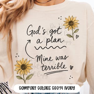 Puede incluir: Una sudadera de manga larga color marfil con un estampado gr&aacute;fico de dos girasoles amarillos y texto negro en cursiva que dice God's got a plan, con una flecha que apunta a Mine was terrible, decorado con peque&ntilde;os corazones y puntos negros.