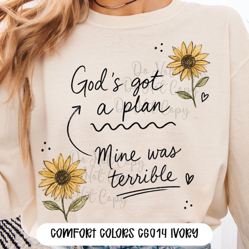 Puede incluir: Una sudadera de manga larga color marfil con un estampado gr&aacute;fico de dos girasoles amarillos y texto negro en cursiva que dice God's got a plan, con una flecha que apunta a Mine was terrible, decorado con peque&ntilde;os corazones y puntos negros.
