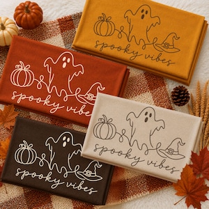 Op de afbeelding: Vier gevouwen stoffen rechthoeken in mosterdgeel, gebrand oranje, donkerbruin en crème. Elk is voorzien van een witte lijntekening van een pompoen, een geest en een heksenhoed met de tekst "spooky vibes" geschreven in een cursief lettertype.