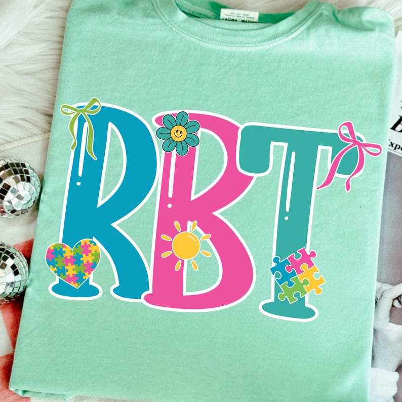 May include: A light green t-shirt with the letters "RBT" in blue and pink, decorated with a flower, a sun, a heart, and puzzle pieces. The shirt has a crew neck and short sleeves.