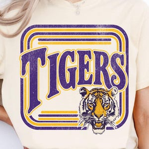 May include: A cream-colored t-shirt featuring a graphic design with the word TIGERS in large purple letters. The text is framed by concentric purple and yellow rounded rectangular lines. A detailed tiger head illustration is positioned below the text.