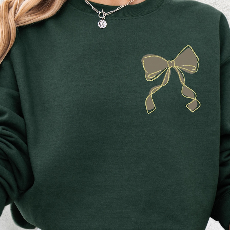 Puede incluir: Sudadera verde oscuro con un dise&ntilde;o de lazo contorneado en dorado en la parte delantera. El lazo est&aacute; relleno de un color marr&oacute;n apagado. La sudadera tiene cuello redondo y mangas largas.