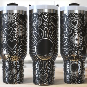 Könnte beinhalten: Drei Edelstahlbecher mit schwarzen Deckeln und Griffen. Jeder Becher hat ein anderes Design mit silbernen Herzen, Blumen und Wirbeln auf schwarzem Hintergrund.