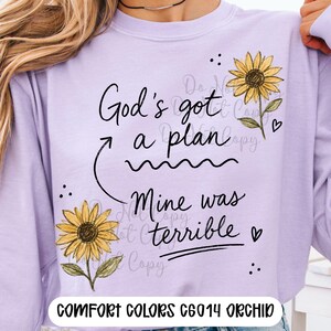Puede incluir: Una sudadera de cuello redondo color morado claro con el texto negro God's got a plan Mine was terrible, una flecha curva, dos girasoles amarillos con hojas verdes, corazones peque&ntilde;os y puntos. El texto Comfort Colors C6014 Orchid es visible.