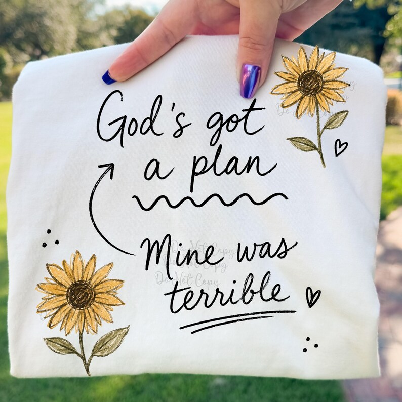 Puede incluir: Una camiseta blanca con texto negro en cursiva que dice God's got a plan, con una flecha que apunta al texto Mine was terrible. El dise&ntilde;o incluye dos girasoles amarillos con hojas verdes y peque&ntilde;os corazones y puntos negros como acentos.