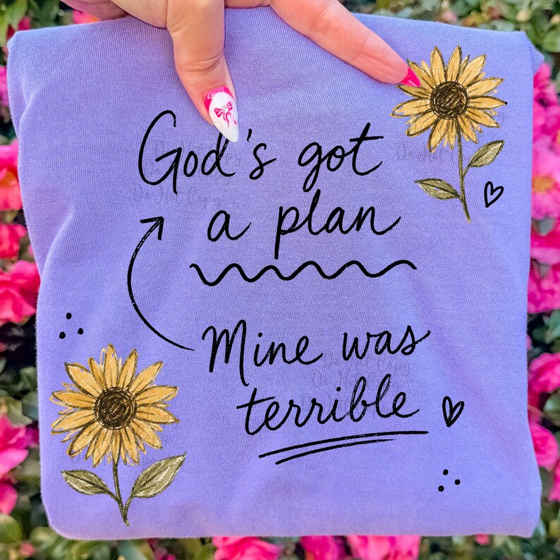 Puede incluir: Una camiseta de color morado claro con el texto en negro "God's got a plan" y "Mine was terrible", junto a una flecha curva que conecta ambas frases. El dise&ntilde;o incluye dos girasoles amarillos con hojas verdes y peque&ntilde;os corazones negros.