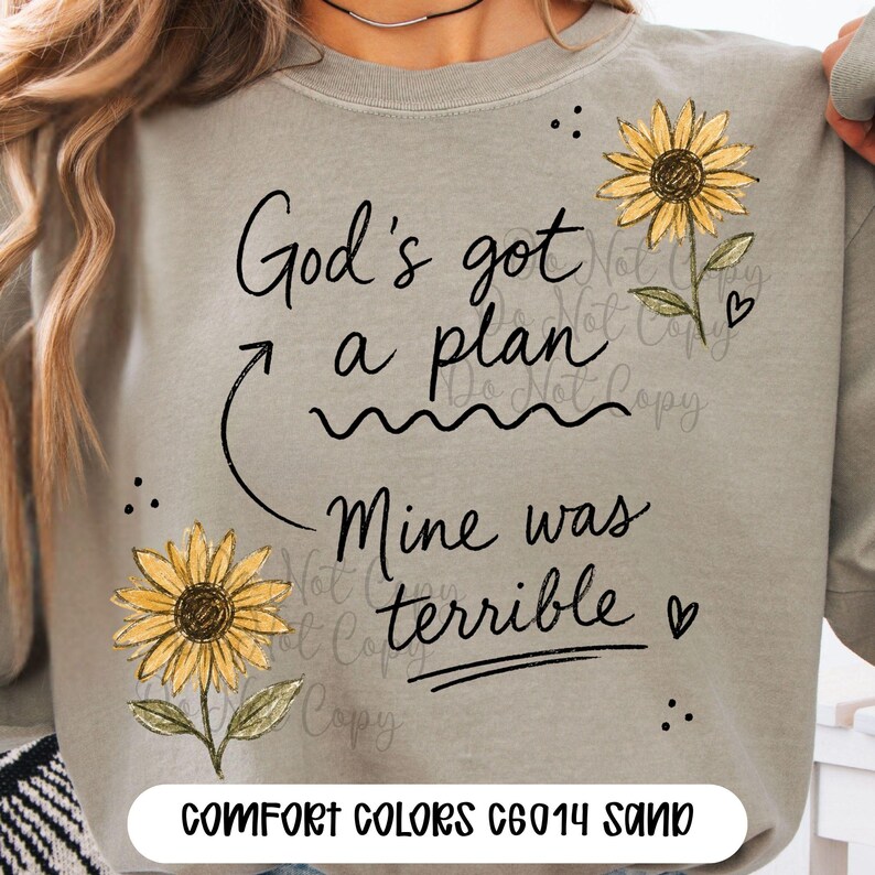 Puede incluir: Una sudadera de cuello redondo color arena con el texto negro God's got a plan, una flecha, una l&iacute;nea ondulada y el texto Mine was terrible. El dise&ntilde;o incluye dos girasoles amarillos con hojas verdes y peque&ntilde;os corazones y puntos negros.