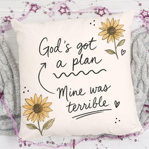 Puede incluir: Un coj&iacute;n cuadrado de color blanco roto con el texto impreso en negro God's got a plan, con una flecha que se&ntilde;ala el texto Mine was terrible. El dise&ntilde;o incluye dos girasoles amarillos con tallos verdes y peque&ntilde;os corazones negros.
