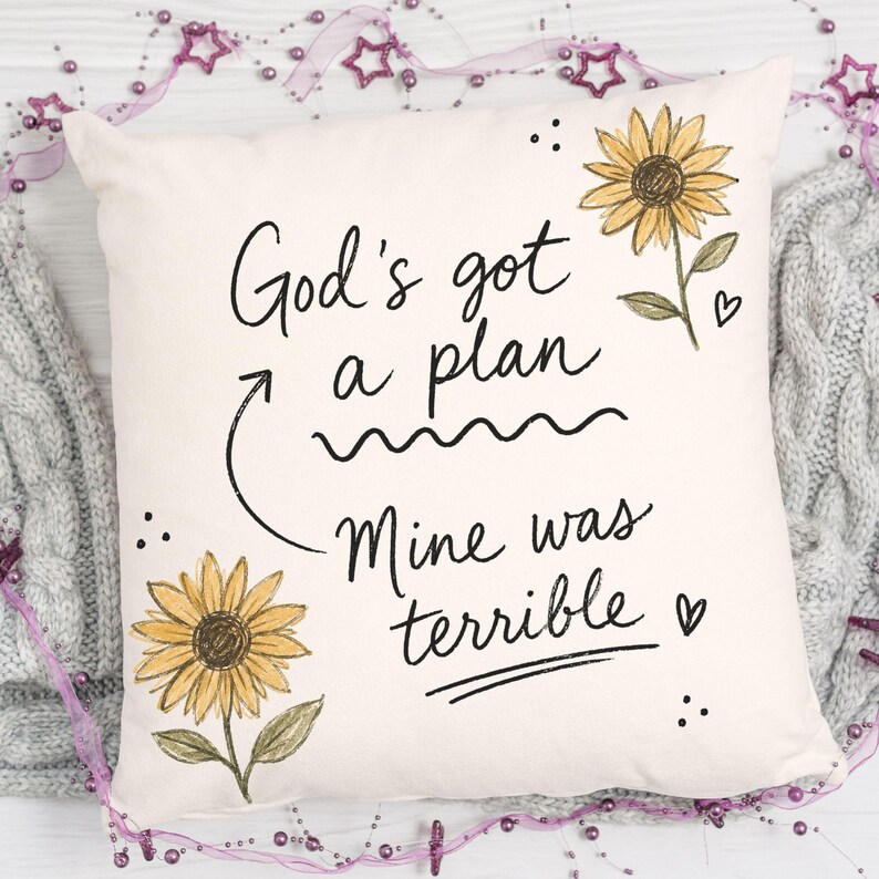 Puede incluir: Un coj&iacute;n cuadrado de color blanco roto con el texto impreso en negro God's got a plan, con una flecha que se&ntilde;ala el texto Mine was terrible. El dise&ntilde;o incluye dos girasoles amarillos con tallos verdes y peque&ntilde;os corazones negros.