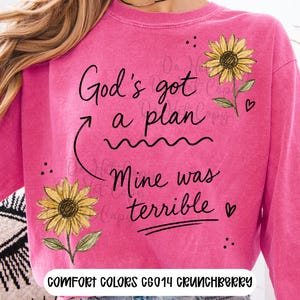 Puede incluir: Una sudadera de color rosa brillante con mangas largas, decorada con dos girasoles amarillos y el texto negro "God's got a plan" con una flecha que se&ntilde;ala "Mine was terrible". La prenda tiene cuello redondo y un aspecto de tela desgastada.