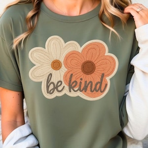 May include: A green t-shirt with a white and orange embroidered flower design and the text "be kind".