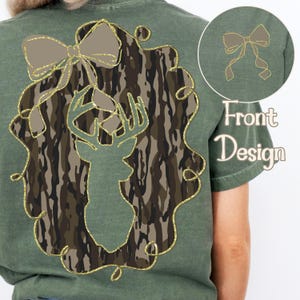 Puede incluir: Camiseta verde con una silueta de cabeza de ciervo en un dise&ntilde;o de camuflaje, enmarcada por un contorno dorado brillante y un lazo. Un dise&ntilde;o de lazo m&aacute;s peque&ntilde;o est&aacute; en la esquina superior derecha. Las palabras "Front Design" est&aacute;n impresas debajo.