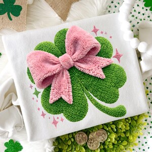 May include: White t-shirt featuring a large green shamrock with a fluffy pink bow in the center. Pink star accents surround the shamrock. The design is likely for St. Patrick's Day or a similar themed event.