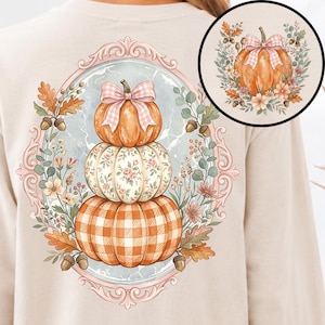May include: A beige long-sleeve shirt featuring a large oval frame graphic on the back. The design shows a stack of three pumpkins decorated with a pink gingham bow, floral patterns, and orange plaid, surrounded by acorns, leaves, and small flowers.