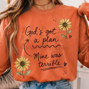 Puede incluir: Una sudadera naranja de cuello redondo con el texto negro God's got a plan Mine was terrible. El dise&ntilde;o incluye dos girasoles amarillos con tallos y hojas verdes, una flecha curva que apunta al texto y peque&ntilde;os iconos de corazones negros.