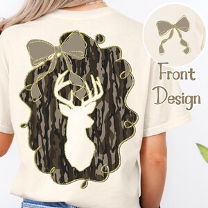 Puede incluir: Camiseta color crema con una silueta de cabeza de ciervo dentro de un dise&ntilde;o de camuflaje, enmarcada por un contorno dorado brillante y un lazo. La parte trasera de la camiseta tiene un peque&ntilde;o dise&ntilde;o de lazo y las palabras "Front Design".