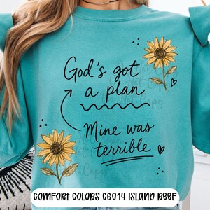 Puede incluir: Una sudadera de cuello redondo color verde azulado con el texto impreso en negro God's got a plan Mine was terrible. El dise&ntilde;o incluye dos girasoles amarillos con tallos y hojas verdes, junto con peque&ntilde;os corazones y puntos negros.