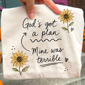 Puede incluir: Una camiseta de color crema con un gr&aacute;fico en letra negra que dice God's got a plan, con una flecha que apunta al texto Mine was terrible. El dise&ntilde;o est&aacute; decorado con dos girasoles amarillos con hojas verdes y peque&ntilde;os corazones negros.