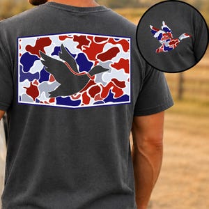 K&ouml;nnte beinhalten: Dunkelgraues T-Shirt mit einem rechteckigen Aufdruck auf dem R&uuml;cken, der eine Entensilhouette zeigt. Die Grafik hat ein rotes, wei&szlig;es und blaues Tarnmuster mit wei&szlig;em Rand. Eine kleinere Entengrafik befindet sich oben rechts.