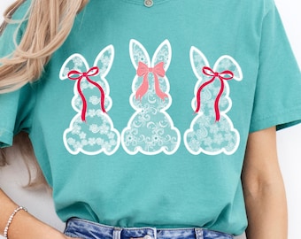 May include: Teal t-shirt featuring three white lace bunny silhouettes. Each bunny has a pink bow.