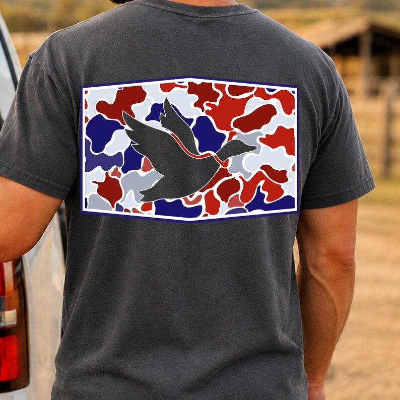 K&ouml;nnte beinhalten: Dunkelgraues T-Shirt mit einer Grafik einer fliegenden Ente. Die Ente ist rot umrandet und vor einem Camouflagemuster aus Rot, Wei&szlig; und Blau platziert. Das Design wird von einem wei&szlig;en Rahmen eingefasst.