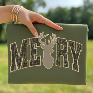 May include: Olive green sweatshirt with the word "MERRY" in a collegiate style font. The letters have a camouflage pattern with gold glitter trim. A deer silhouette is centered between the words.
