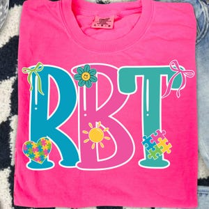 May include: A bright pink t-shirt with the letters "RBT" in teal and pink. The design includes a flower, a sun, a heart, and a puzzle piece, all in vibrant colors. The shirt is made of a soft material.