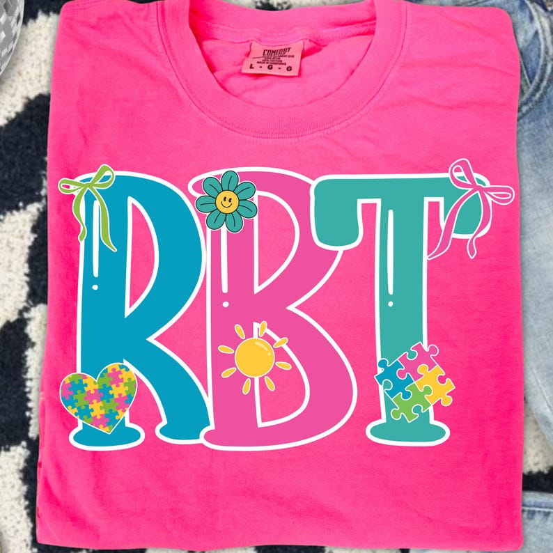 May include: A bright pink t-shirt with the letters "RBT" in teal and pink. The design includes a flower, a sun, a heart, and a puzzle piece, all in vibrant colors. The shirt is made of a soft material.