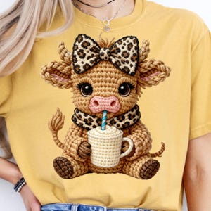 May include: A yellow t-shirt featuring a crocheted cow illustration. The cow has a bow, scarf, and is holding a mug with a straw. The cow is brown, with a pink nose and large eyes. The bow and scarf have a leopard print design.