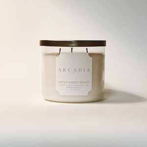 May include: A clear glass candle jar with a dark brown lid. The candle is filled with a creamy wax and has a label that reads "Arcadia" and "Fresh Baked Bread". The candle weighs 15.5 oz.