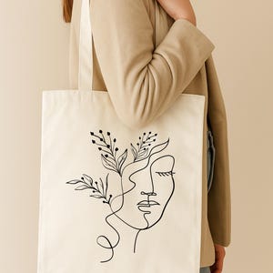 Könnte beinhalten: Cremefarbene Tragetasche mit einer schwarzen Strichzeichnung eines Gesichts und floralen Elementen. Die Tasche hat ein schlichtes, minimalistisches Design mit einem langen Riemen. Die Tragetasche ist aus robustem Stoff gefertigt.