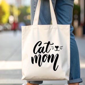 Könnte beinhalten: Eine weiße Stofftasche mit dem schwarzen Schriftzug "Cat mom" und einer Katzenkopf-Grafik. Die Tasche wird von einer Person getragen, die blaue Jeans und weiße Turnschuhe trägt.