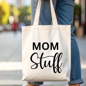 Op de afbeelding: Een witte canvas tas met de zwarte tekst "MOM Stuff". De tas wordt vastgehouden door iemand die een blauwe spijkerbroek en witte sneakers draagt. De achtergrond is wazig.