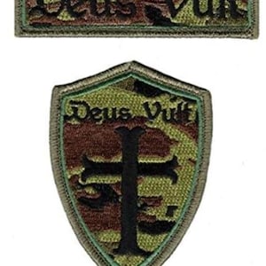 Puede incluir: Dos parches bordados con fondo de camuflaje. El primer parche es rectangular y dice "Deus Vult". El segundo parche tiene forma de escudo y tiene una cruz negra en el centro. El escudo también dice "Deus Vult".