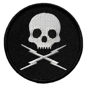 Puede incluir: Parche redondo bordado en negro con una calavera blanca y rayos cruzados. La calavera tiene cuencas oscuras y una textura detallada. Los rayos blancos se cruzan. Objeto decorativo.