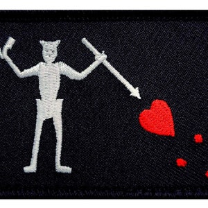 May include: A black embroidered patch with a white stick figure holding a spear that is piercing a red heart. There are three red drops of blood below the heart.