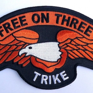 Puede incluir: Parche bordado negro con una pancarta naranja que dice "FREE ON THREE TRIKE" y un águila blanca con alas naranjas.