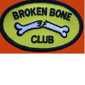 Könnte beinhalten: Ein gelber ovaler Patch mit schwarzen Nähten an den Rändern. Der Patch hat einen weißen gebrochenen Knochen mit schwarzem Umriss und den Text "BROKEN BONE CLUB" in Schwarz.