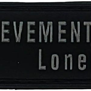 Puede incluir: Parche rectangular negro con bordes redondeados. El texto "ACHIEVEMENT EARNED" y "Loner" está en letras grises. Un gráfico verde y gris de un candado está en el lado izquierdo.