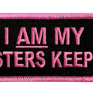 May include: Pink embroidered patch with black background. The text reads "I AM MY SISTERS KEEPER".