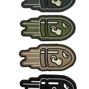May include: Four different coloured patches featuring a cartoon bullet with a face and a fist. The bullet is black, green, brown, and black.
