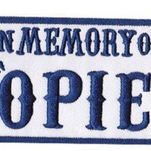Op de afbeelding: Blauw en wit geborduurd patch met de tekst "In Memory of 'Opie'".