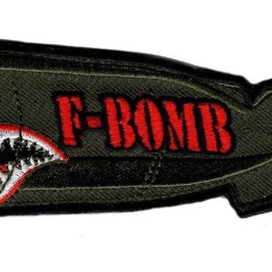 May include: Embroidered patch in the shape of a bomb, with a shark mouth design and the text "F-BOMB" in red. The patch is olive green with a yellow radiation symbol.