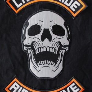 May include: Two black and orange embroidered patches with white text. The top patch reads "Live to Ride" and the bottom patch reads "Ride to Live". A white skull with black eyes and an open mouth is centered between the patches.
